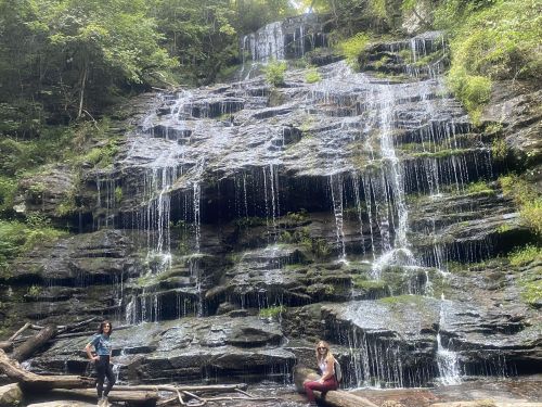 Two people sit near a waterfall surrounded by lush greenery and rocky terrain. Water cascades down the rocks.