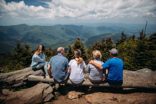A group of five people sitting on a log, overlooking a scenic mountain view under a cloudy sky.