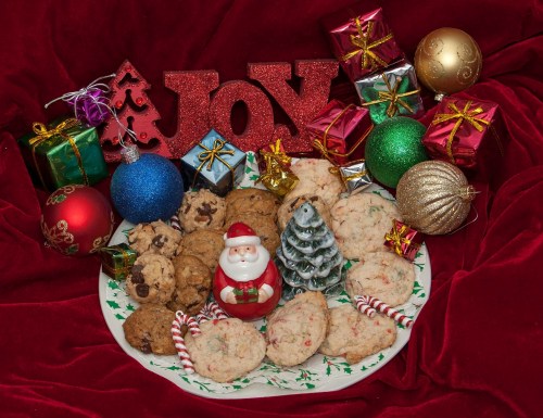 A festive plate of cookies surrounded by colorful ornaments, gifts, and a Santa figurine on a red background.