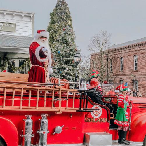 Santa Claus waves from a festive red fire truck, surrounded by Christmas trees and cheerful elves.