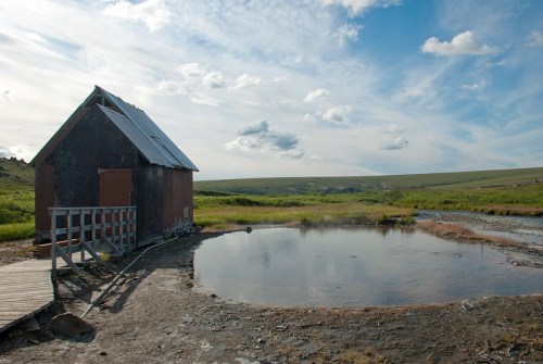 A rustic shed beside a small pond, surrounded by green fields and a blue sky with scattered clouds.