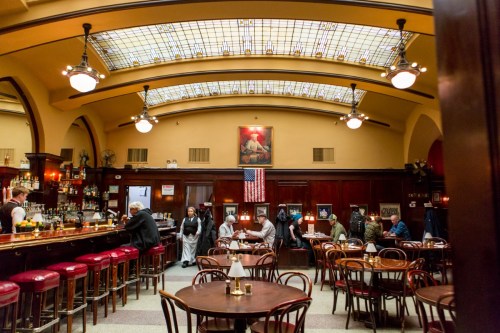 A cozy restaurant interior with a bar, wooden tables, and patrons enjoying their meals under a glass ceiling.
