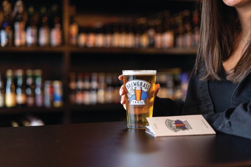 A person holding a pint of beer with a Brewgrass Trail logo, sitting at a bar with shelves of bottles in the background.