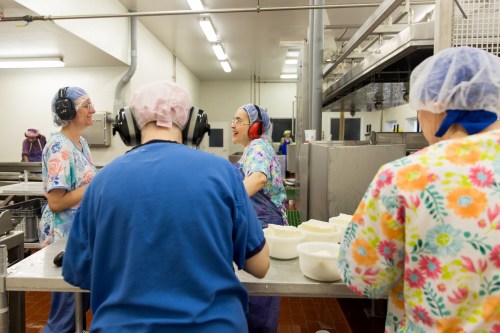 Four people in colorful scrubs and hairnets work in a kitchen, smiling and chatting near a stainless steel counter.