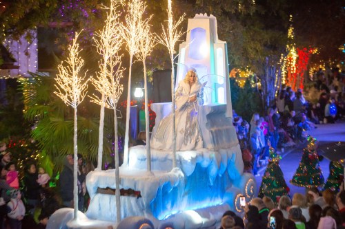 A festive parade scene featuring a woman in a white gown on a float with icy decorations and illuminated trees.