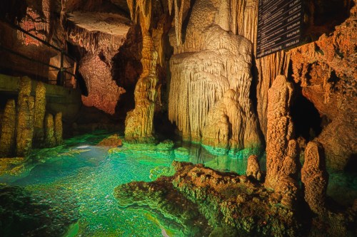 A stunning underground cave with stalactites and stalagmites, featuring a shimmering turquoise pool of water.