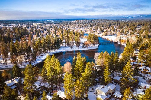 Aerial view of a snowy landscape with a river, trees, and houses in a scenic, winter setting.