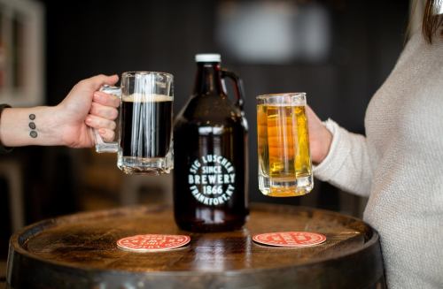 Two people clink beer mugs over a wooden table, featuring a growler and coasters from a brewery.