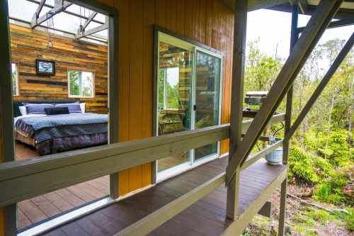 A cozy cabin interior with a wooden bed and large windows, surrounded by lush greenery outside.