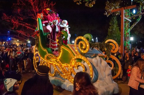 A festive parade scene with Santa on a decorated sleigh, surrounded by a cheering crowd and colorful lights.