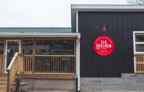 Exterior of Sig Luscher Brewery, featuring a wooden deck, black siding, and a red circular sign with the brewery's name.
