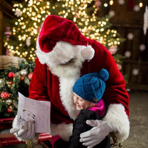 Santa Claus holds a letter while joyfully interacting with a young girl in a festive, decorated setting.