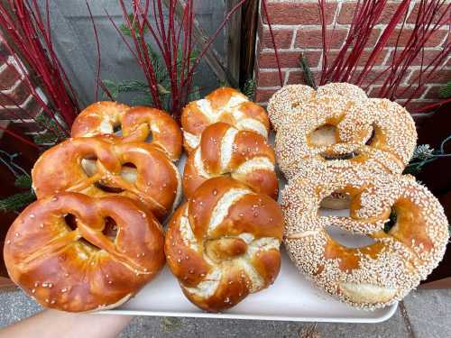 A tray of freshly baked pretzels, some plain and some topped with sesame seeds, set against a brick background.