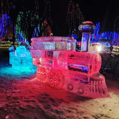 An illuminated ice sculpture of a train, surrounded by colorful lights in a winter setting.