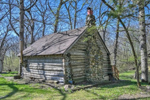 A rustic log cabin with a stone chimney, surrounded by bare trees and greenery on a sunny day.