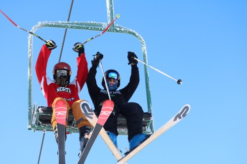 Two skiers on a ski lift, smiling and raising their poles against a clear blue sky. Snow-covered lift structure in background.