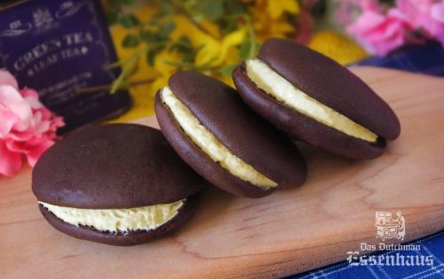 Three chocolate whoopie pies with creamy filling, placed on a wooden board, surrounded by colorful flowers.