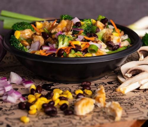 A colorful salad with broccoli, carrots, black beans, corn, and chicken in a black bowl, surrounded by fresh vegetables.