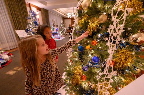 Two children admire a decorated Christmas tree, pointing at ornaments and tinsel in a festive indoor setting.