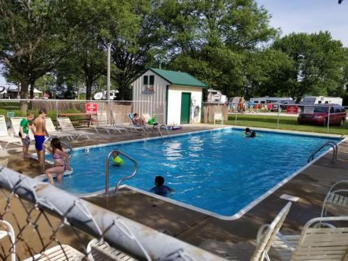 A sunny pool scene with children swimming, lounging chairs, and a small building nearby, surrounded by trees and RVs.