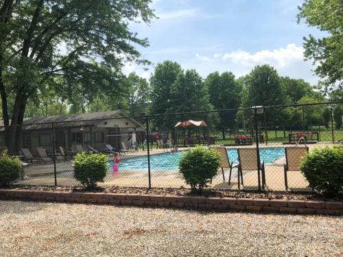 A fenced pool area with lounge chairs, surrounded by trees and a playground in the background on a sunny day.