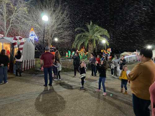 A festive scene with families enjoying holiday lights and artificial snow in a park at night.