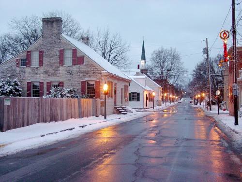 A snowy street scene with historic buildings, glowing street lamps, and a church steeple in the background.