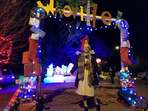 A festive archway decorated with lights and gifts, featuring a person in a colorful outfit and clown wig, celebrating the holiday.