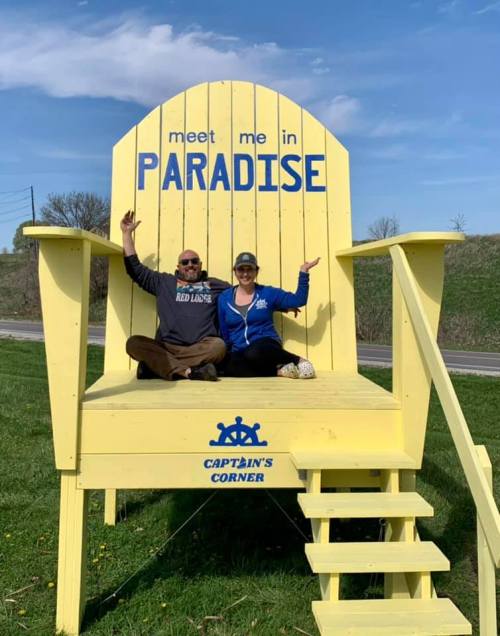 A large yellow chair with "meet me in PARADISE" on it, with two people sitting and posing happily.