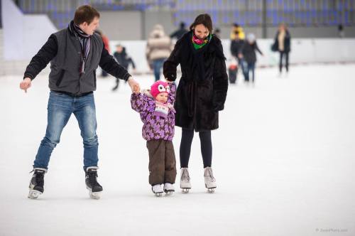A man and woman skate on ice while holding hands with a young girl in a colorful winter outfit.