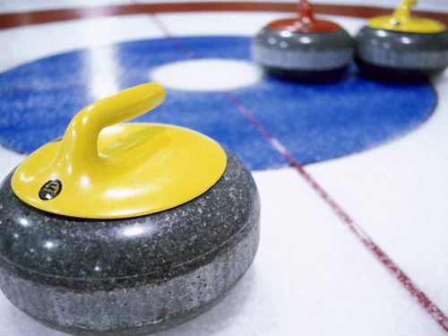 A close-up of a yellow curling stone on an ice rink, with red and gray stones in the background.