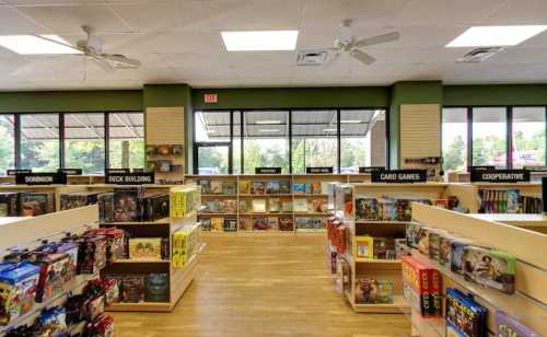 Interior of a game store featuring shelves of board games, card games, and cooperative games, with large windows.