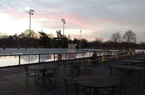 An outdoor ice skating rink at dusk, with skaters gliding on the ice and tables set up nearby under string lights.