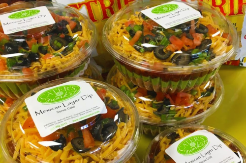 Containers of Mexican layer dip topped with olives and tomatoes, displayed alongside bags of tortilla chips.