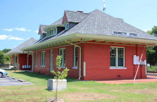 Historic red brick building with a gabled roof, labeled "Mammoth Spring," surrounded by green grass and trees.