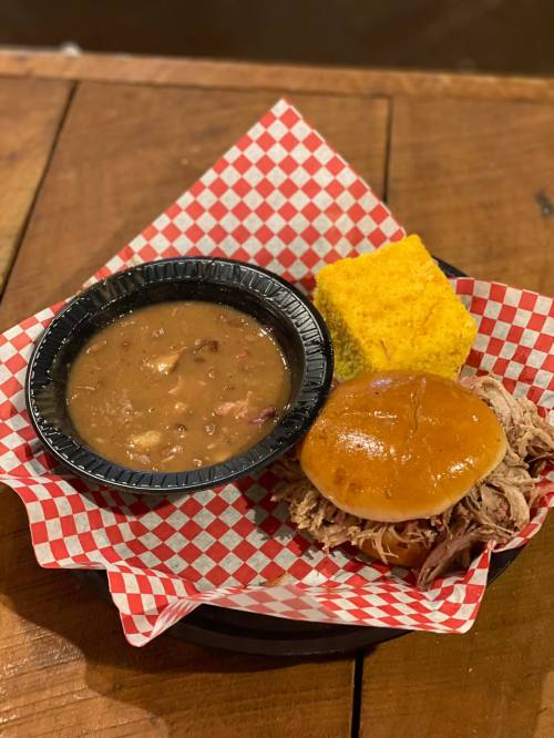 A plate with pulled pork on a bun, cornbread, and a side of beans, served in a checkered basket.