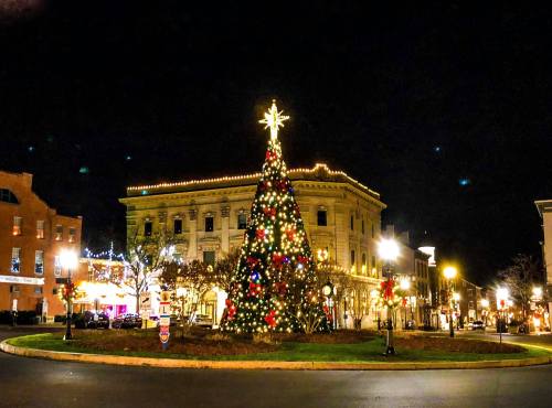 A brightly lit Christmas tree adorned with colorful lights stands in a town square at night, surrounded by festive decorations.