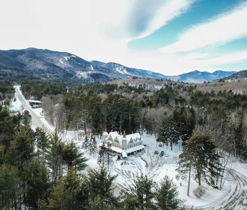 A snowy landscape featuring a white house surrounded by trees, with mountains in the background under a blue sky.