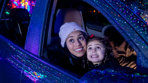 A woman and a girl smile from a car, surrounded by colorful holiday lights reflecting on the window.