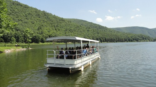 A boat with passengers glides across a calm lake, surrounded by lush green hills under a clear blue sky.