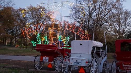 Colorful holiday lights display with "Happy Holidays" sign and decorated horse-drawn carriages in a park setting.