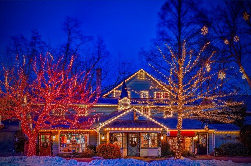 A beautifully decorated house with colorful holiday lights against a twilight sky, surrounded by snow and trees.