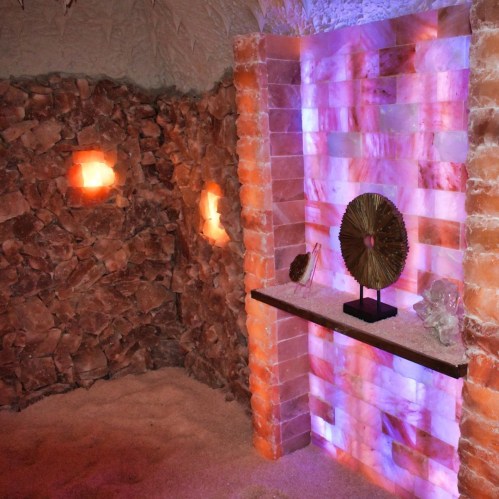 A serene salt room with pink salt walls, glowing salt lamps, and a decorative display on a black shelf.
