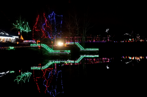 Colorful holiday lights illuminate trees and pathways by a calm lake, reflecting beautifully in the water at night.