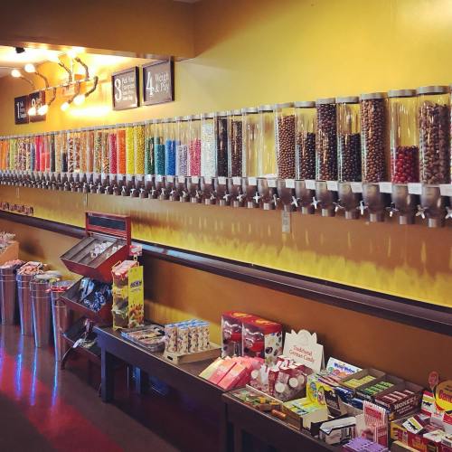A colorful candy wall with various jars and dispensers, alongside shelves of candy boxes and bags on a bright yellow background.