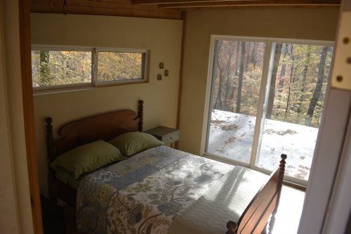 Cozy bedroom with a wooden bed, green pillows, and large windows overlooking a forested area.