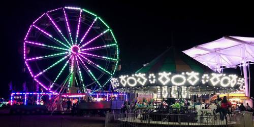 A brightly lit ferris wheel and carousel at night, surrounded by festive lights and people enjoying the fair.