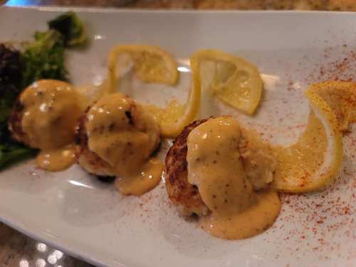 Three crab cakes drizzled with a creamy sauce, garnished with lemon slices and a sprinkle of paprika.