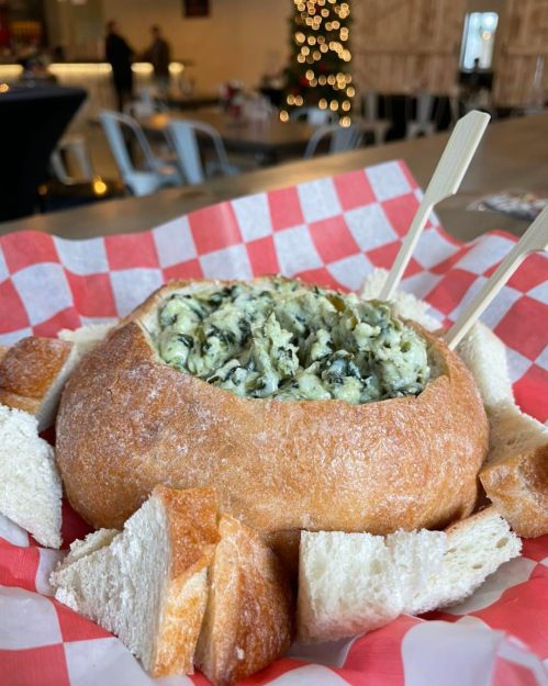 A bread bowl filled with creamy spinach dip, surrounded by pieces of bread, on a red and white checkered paper.