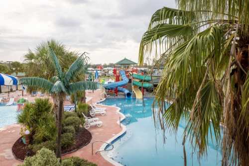 A resort pool area featuring water slides, palm trees, and lounge chairs under a cloudy sky.